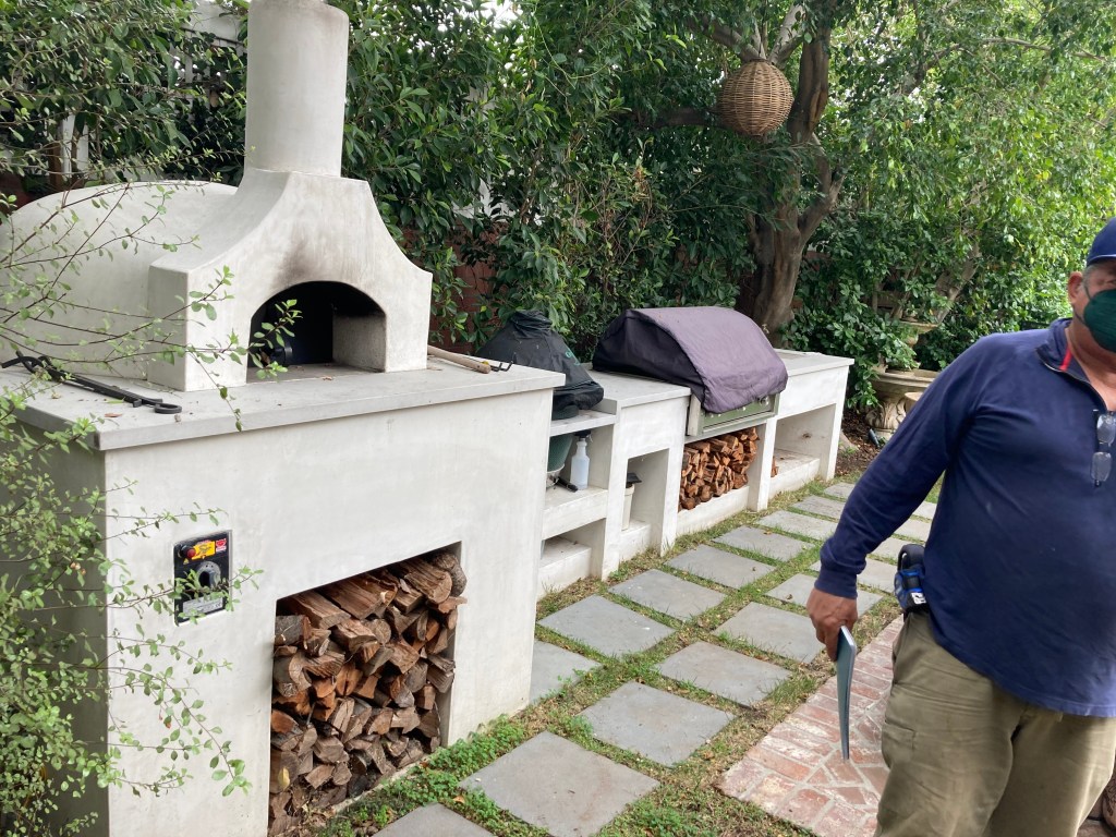 Outdoor cooking area featuring a built-in stone oven, wood storage, and a person standing next to it, surrounded by greenery.