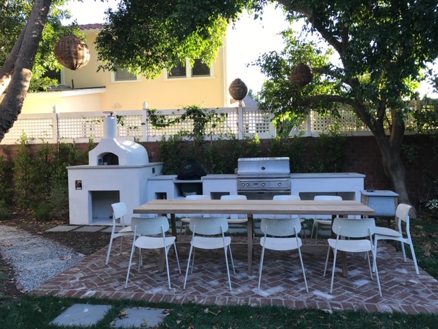 Outdoor kitchen area featuring a stone pizza oven, grill, and a long wooden dining table with white chairs, surrounded by greenery.